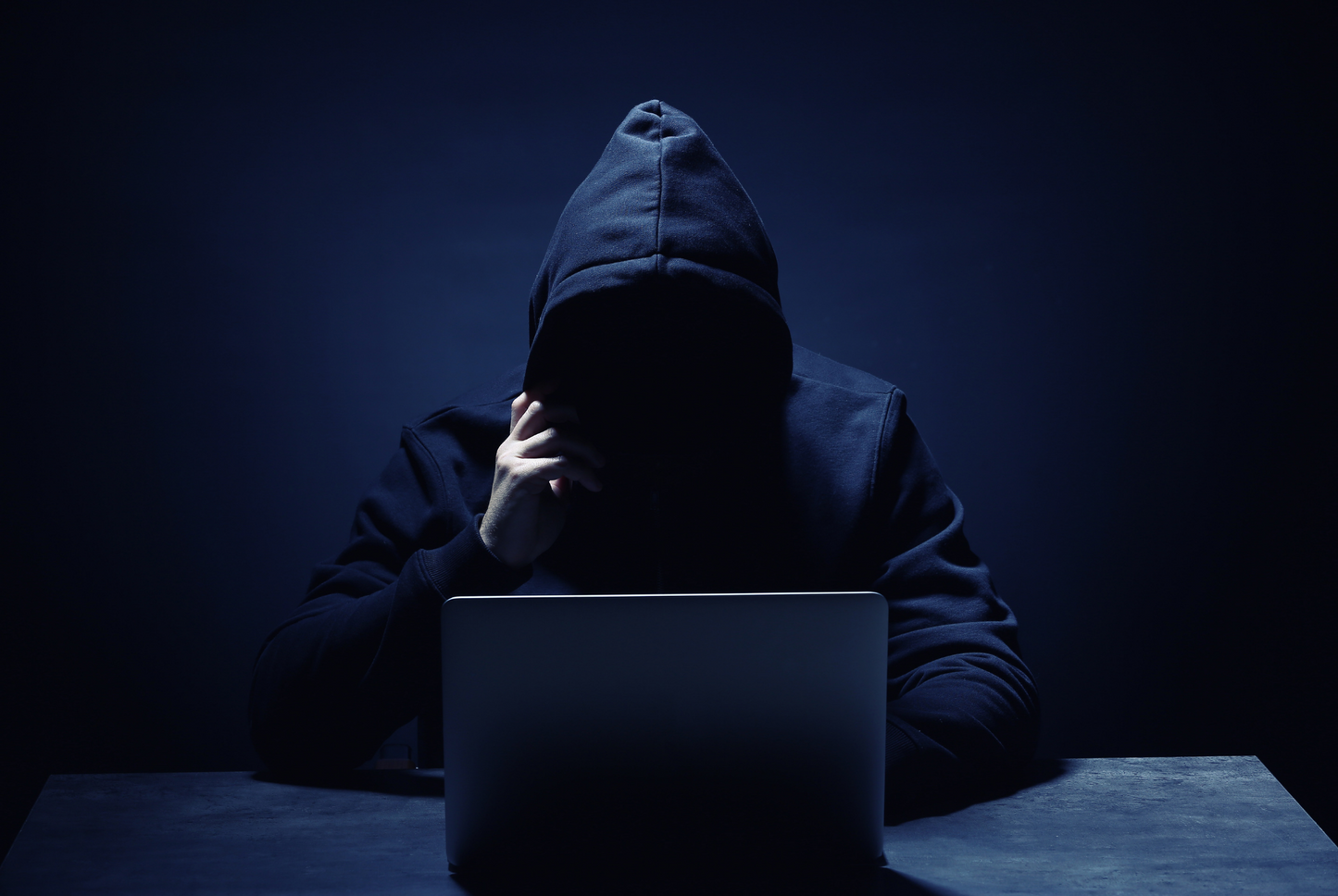 Person wearing a hooded jacket sitting at a table with a laptop in a dark room